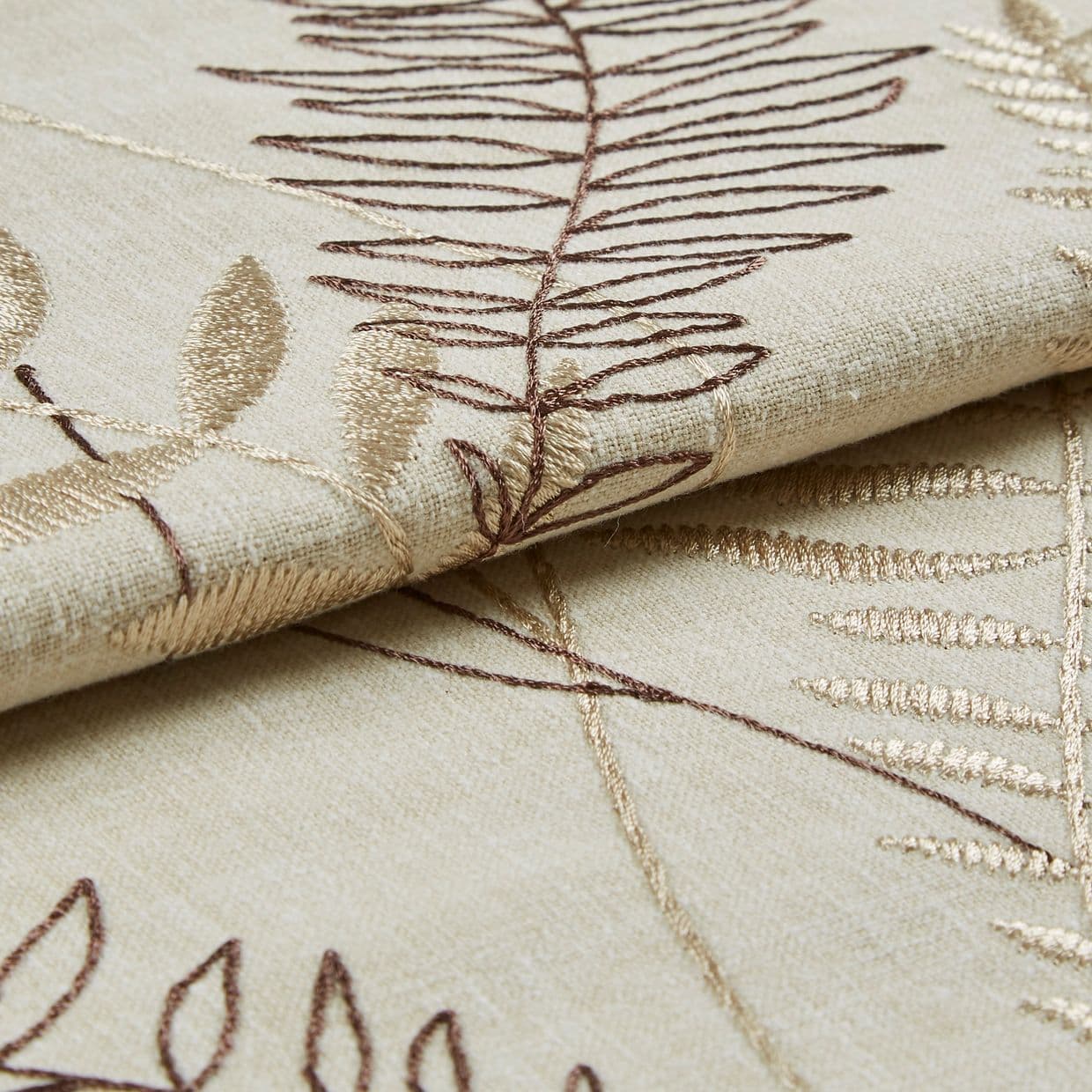 Beige embroidered curtain fabric, folded to show overlapping panels with brown and gold leaf-stitch motifs; embroidery raised and textured; close-up studio shot with soft, even lighting on a neutral surface.