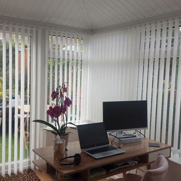 White vertical blinds covering wraparound conservatory windows, narrow slats partially open, filtering daylight; in a bright home office with wooden desk, laptop, monitor, orchid, and garden visible beyond.