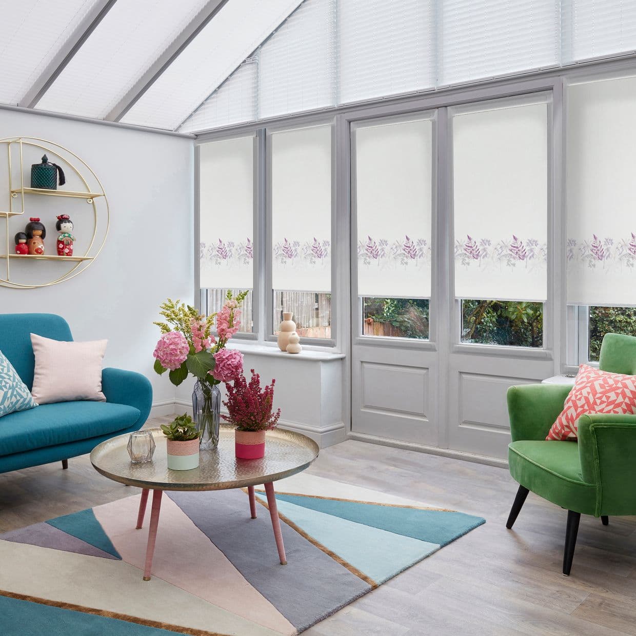 White roller blinds with lavender floral border lowered over tall windows and door, filtering daylight and obscuring view; conservatory with teal sofa, green armchair, pastel rug.