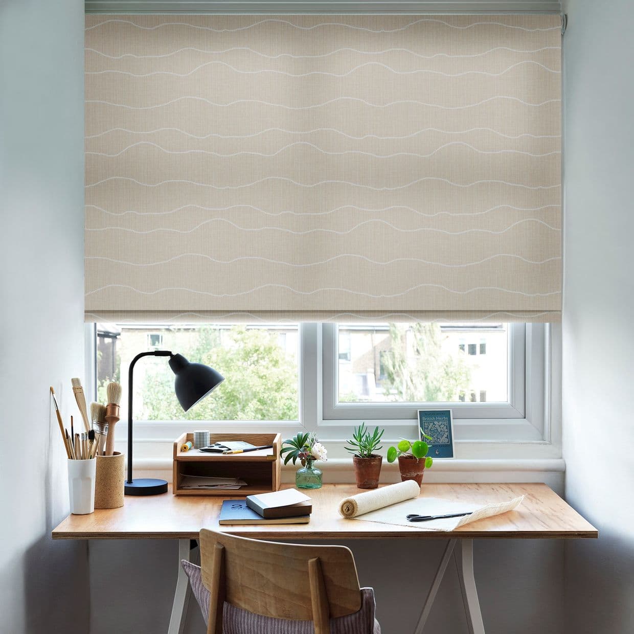 Beige roller blind with subtle white wavy lines lowered across the window, filtering daylight; above a wooden study desk with lamp, brushes, notebooks and potted succulents, view of trees.