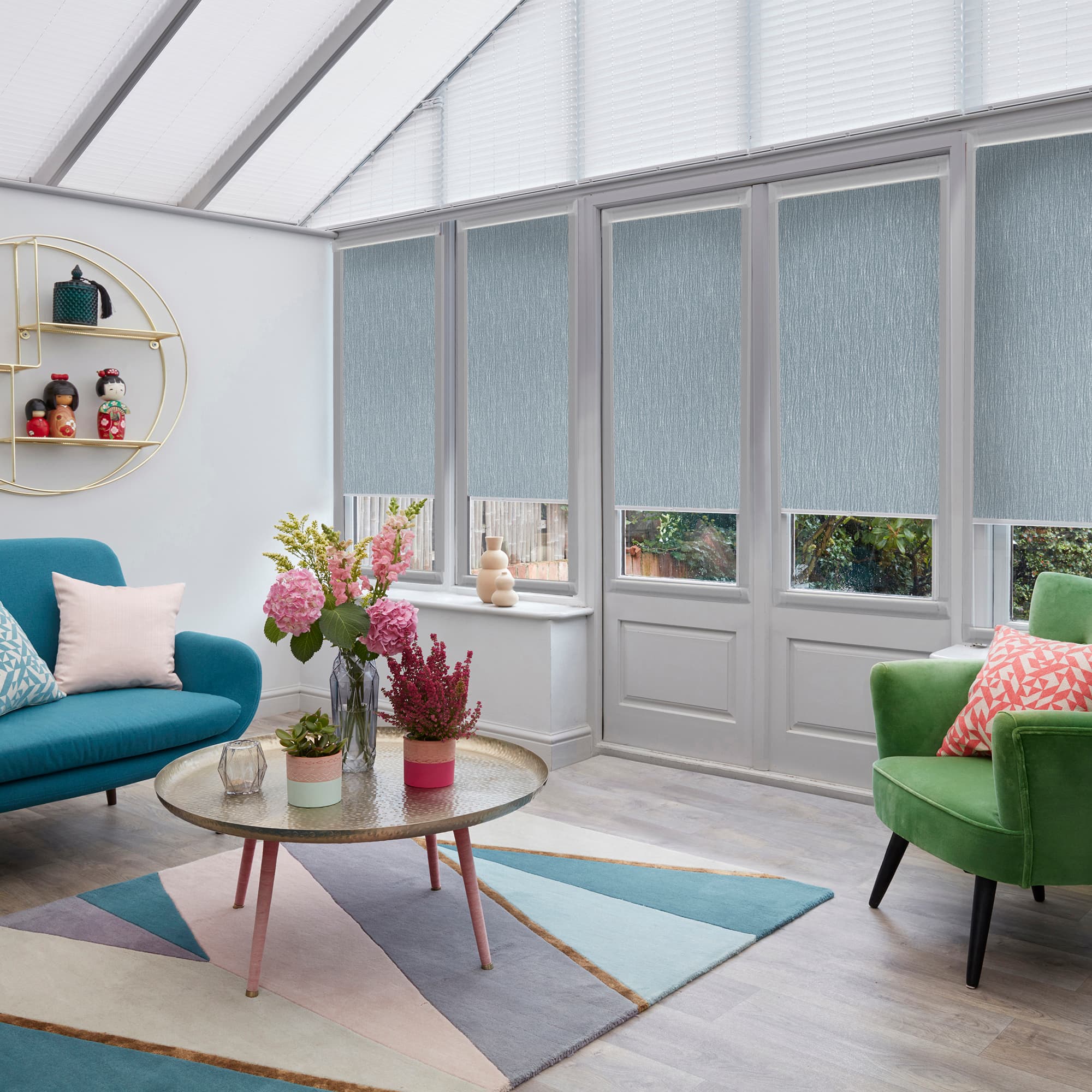Light-blue textured roller blinds covering tall conservatory windows and doors, mostly lowered with narrow bottom openings, filtering daylight; bright living room context with skylight, teal sofa, green armchair, floral centerpiece.