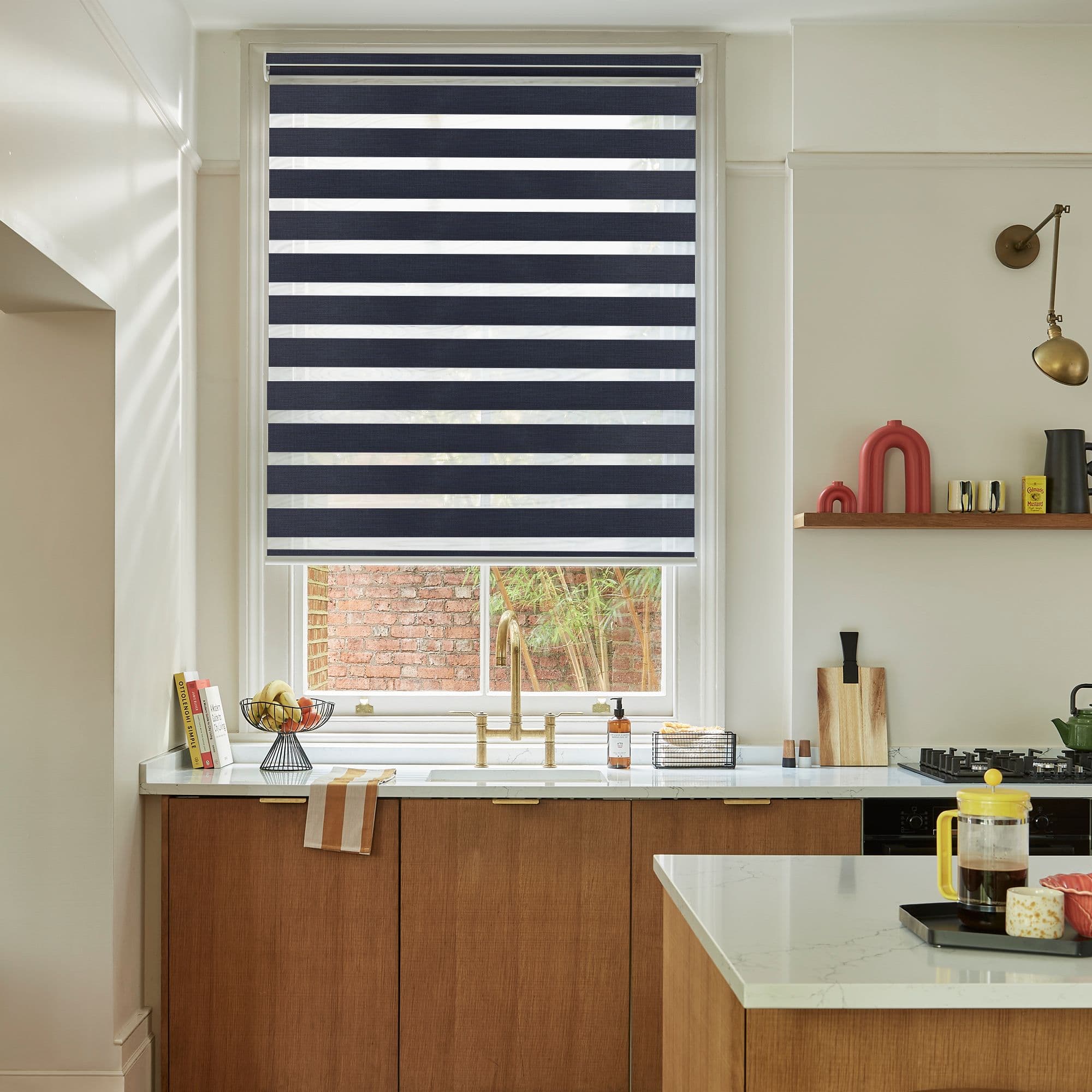 A navy-and-white striped roller blind lowered, alternating opaque and sheer bands filtering light and partially obscuring the view, above a kitchen sink with brass faucet and wood cabinets.