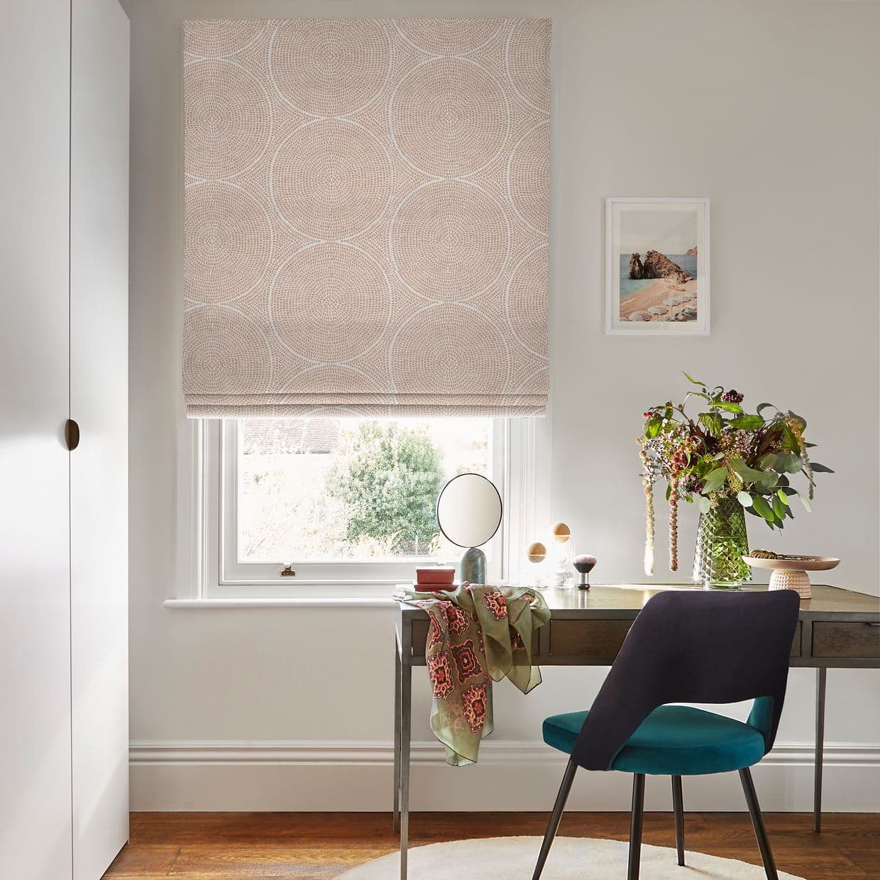 A beige Roman blind with white concentric-circle pattern, partially lowered and folded, filtering daylight through a sash window; small desk/vanity beneath with teal chair, mirror, vase of flowers, framed print.