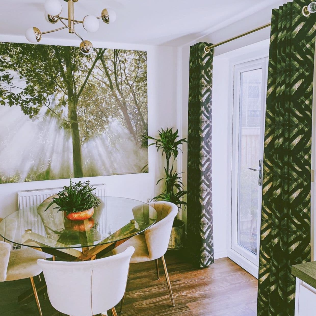 Green patterned grommet curtains flank a glass patio door, pulled aside to admit light; in a dining area with round glass table, upholstered chairs, potted plant, and forest wall mural.