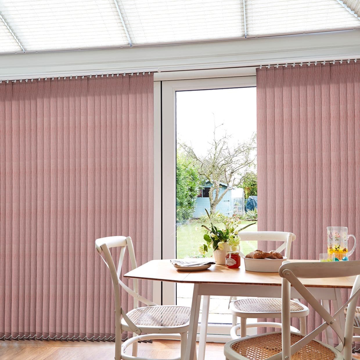 Pink fabric vertical blinds mounted at the ceiling, pulled aside to form an opening and filter daylight. Dining table with chairs, vase and pastries; sunlit backyard with tree and shed.