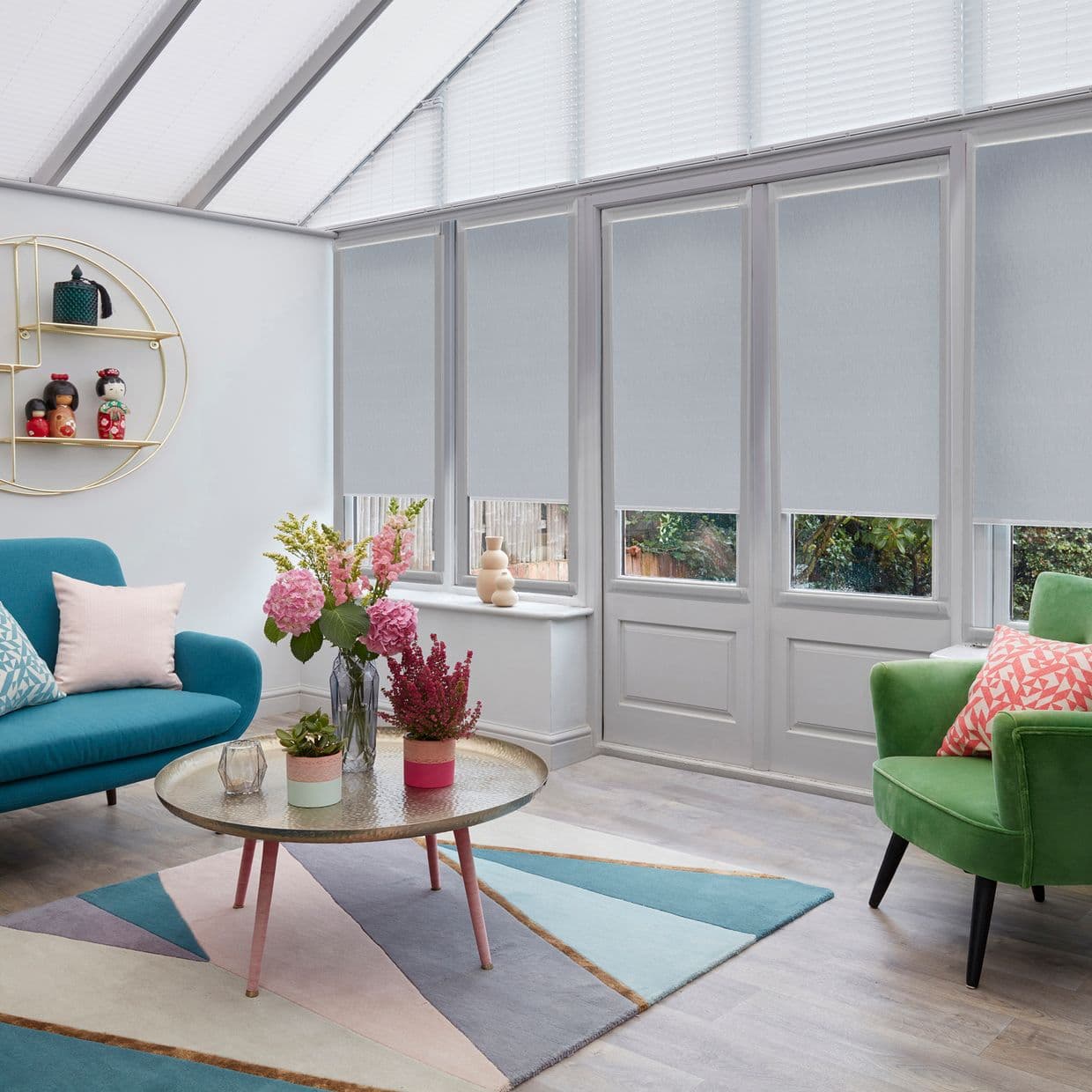 White roller blinds cover tall conservatory windows and glazed roof, lowered halfway filtering soft daylight; living area with pastel sofa, green armchair, geometric rug and coffee table with flowers.