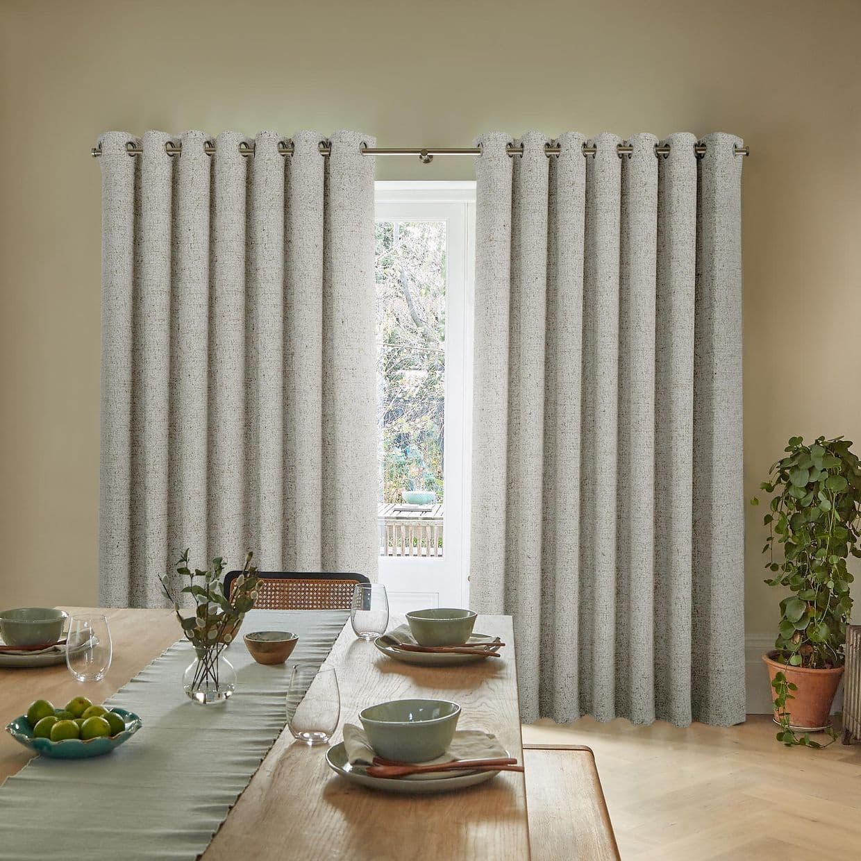 Grommet-top gray curtains, hanging on a metal rod, drawn into pleats with a narrow central gap, filtering daylight in a sunlit dining room with wooden table and potted plant.