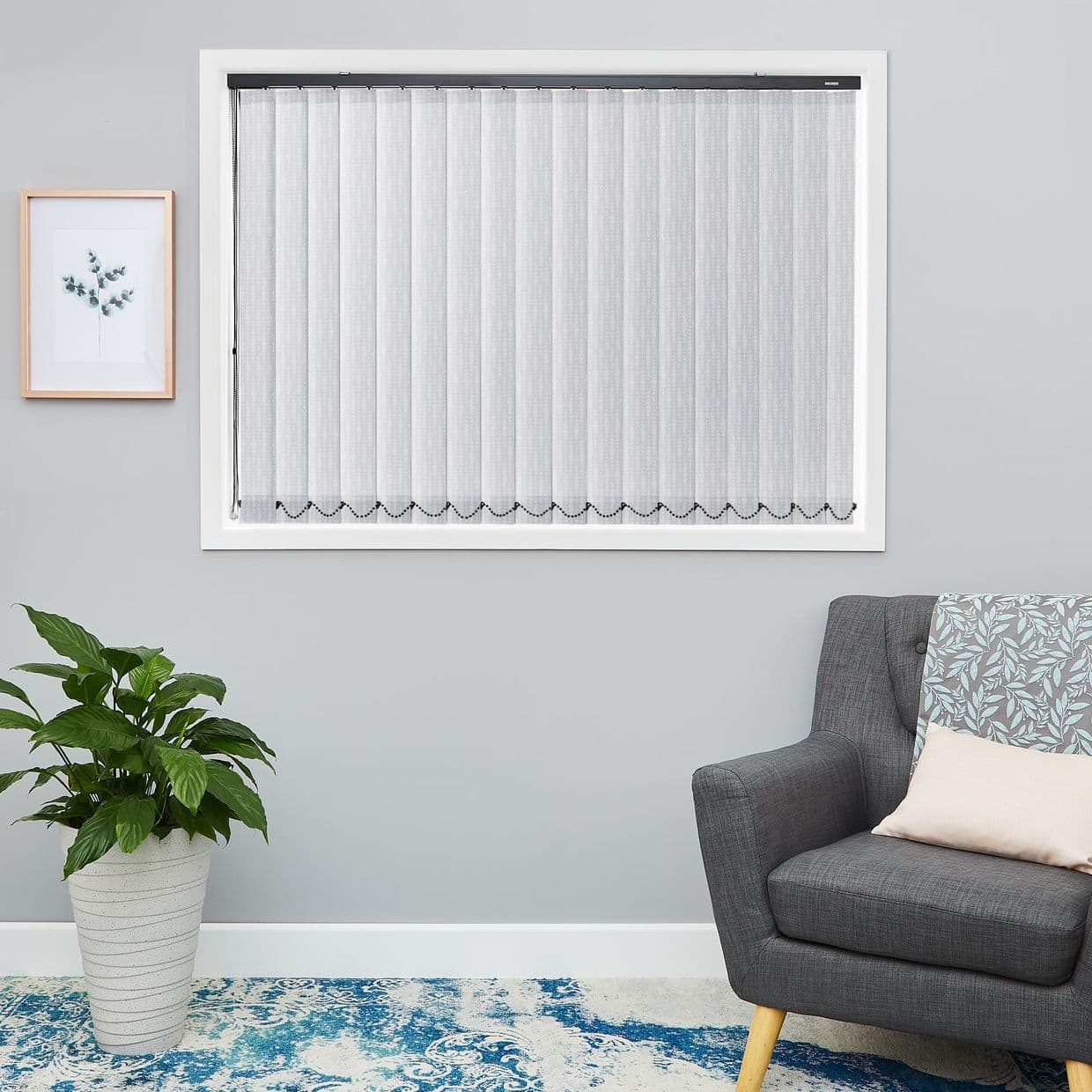 Sheer white vertical blinds covering the window, scalloped beaded hem visible, closed and filtering daylight; living room with grey armchair, potted plant, and framed botanical print.
