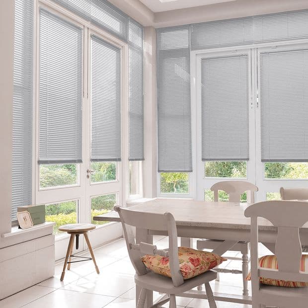 Horizontal Venetian blinds cover tall French windows and doors, lowered and partially closed, filtering soft daylight into a bright dining area with white table, chairs, and a green garden view.