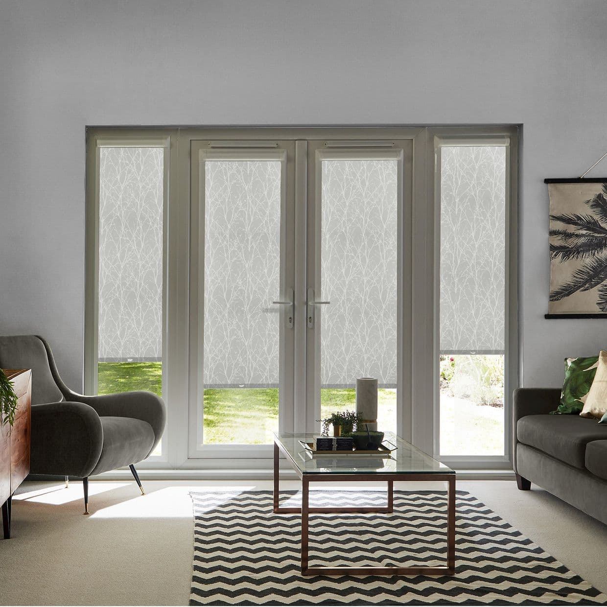 White roller blinds with a subtle branch pattern covering four-panel French doors and side windows, lowered and filtering daylight (small bottom gaps reveal lawn) â€” modern living room, soft natural light.