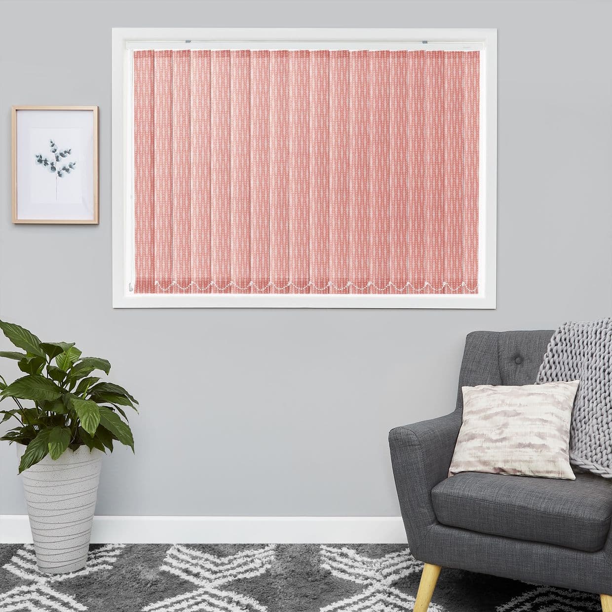 Pink patterned vertical blinds covering a white-framed window, closed and filtering daylight. On a pale gray living-room wall with framed botanical print, potted plant, gray armchair, throw and rug.