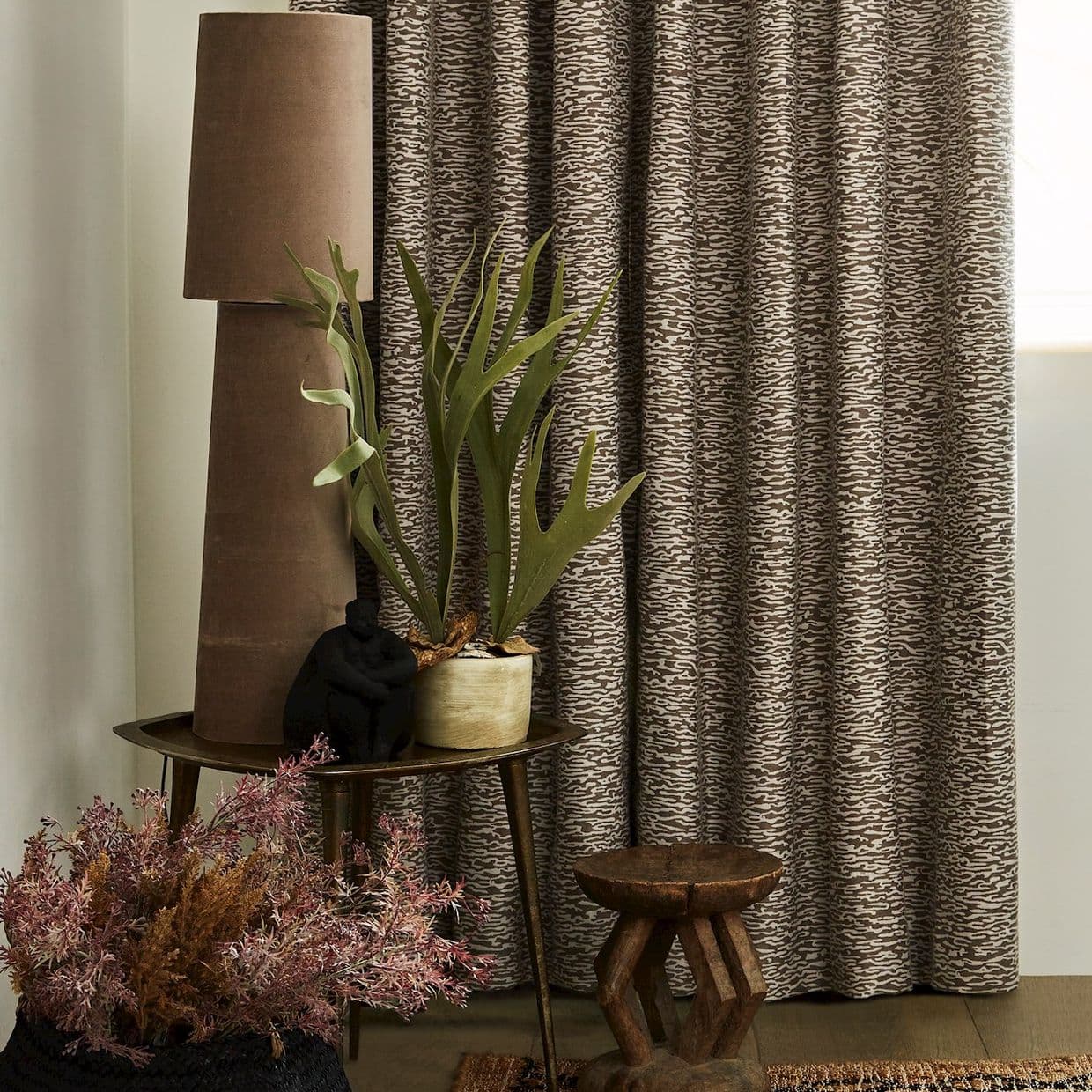 Patterned brown curtains (full-length, heavy fabric) drawn closed, softly filtering light; beside them a side table holds a tall lamp, potted staghorn fern and decorative objects in a warm corner.