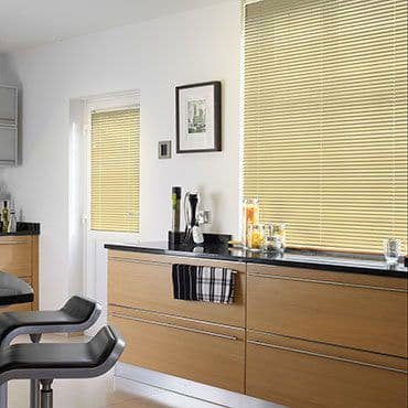 Beige horizontal Venetian blinds covering two windows and a glass door, closed and softly filtering daylight, in a modern kitchen with light wood cabinets, black countertop, stools, and countertop accessories.