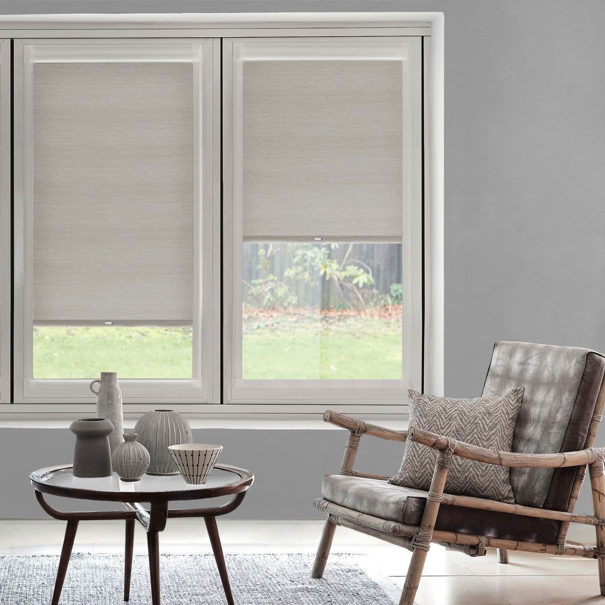 Light-gray fabric roller shades covering twin windows, textured with visible hem bar; partially lowered, filtering daylight into a neutral living room with rattan armchair and backyard view.