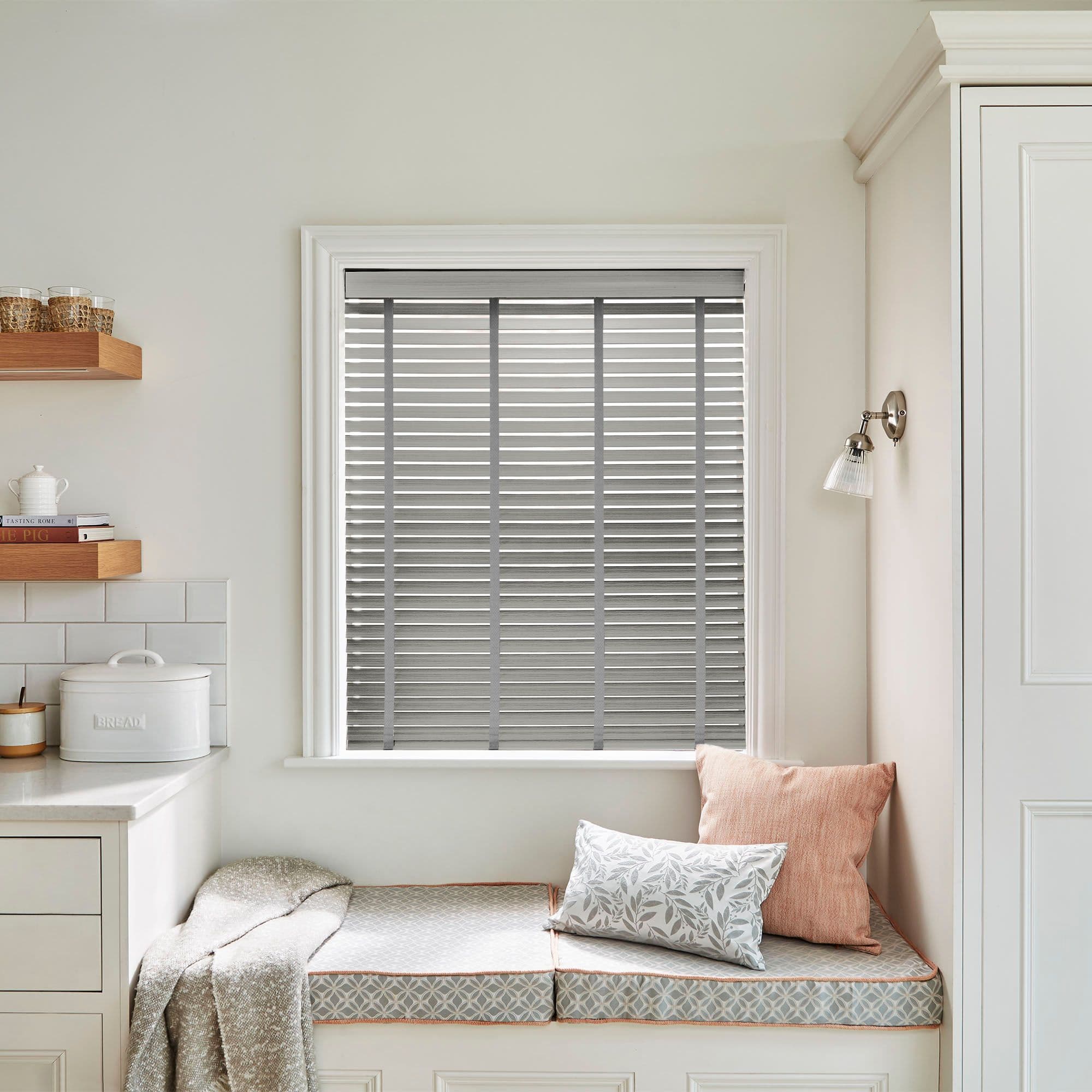 Grey horizontal blinds with vertical ladder tapes covering the window, closed and filtering soft daylight; cozy kitchen nook with built-in cushioned bench, pillows, wall sconce, countertop and bread tin.

BREAD