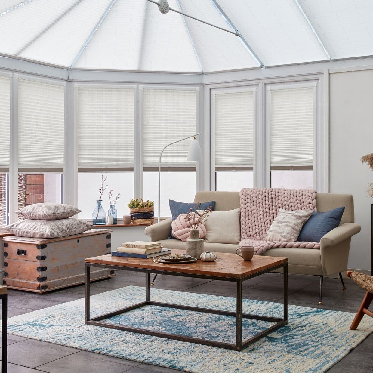White cellular pleated blinds lowered halfway across tall bay windows and conservatory roof, filtering soft daylight into a cozy sunroom with a pale sofa, wooden coffee table, chunky pink throw.