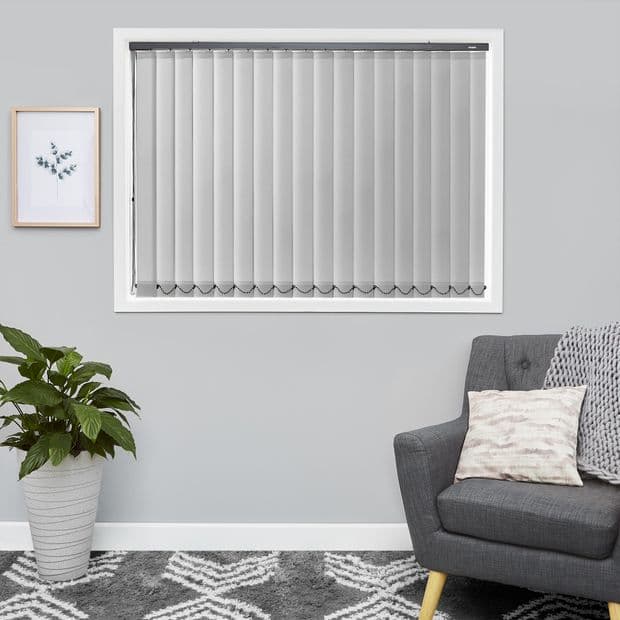 Vertical light-gray blinds covering a rectangular window, slim slats with bottom chain; closed and softly diffusing daylight; in a grey-walled living room with armchair, potted plant, framed art, rug.