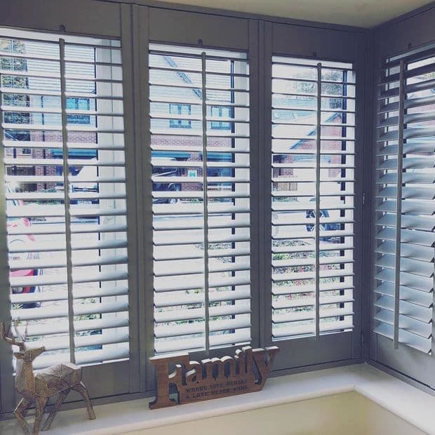 White wooden plantation shutters cover a three-panel bay window, slats tilted open to filter bright daylight and show a suburban street; windowsill holds a wooden "Family" sign and deer ornament.

Transcription:
Family
WHERE LIFE BEGINS & LOVE NEVER ENDS