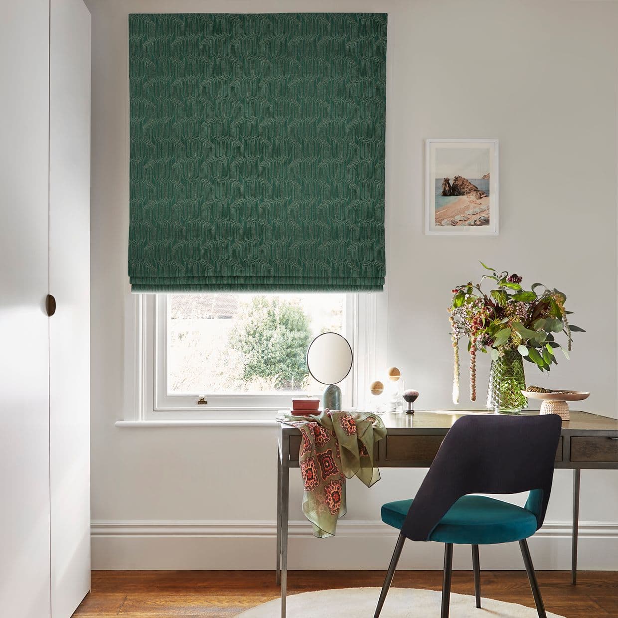 Green patterned Roman blind, lowered and closed, filtering daylight through the window; above a wooden desk with teal chair, vanity mirror, vase of flowers, neutral walls and framed seaside print.