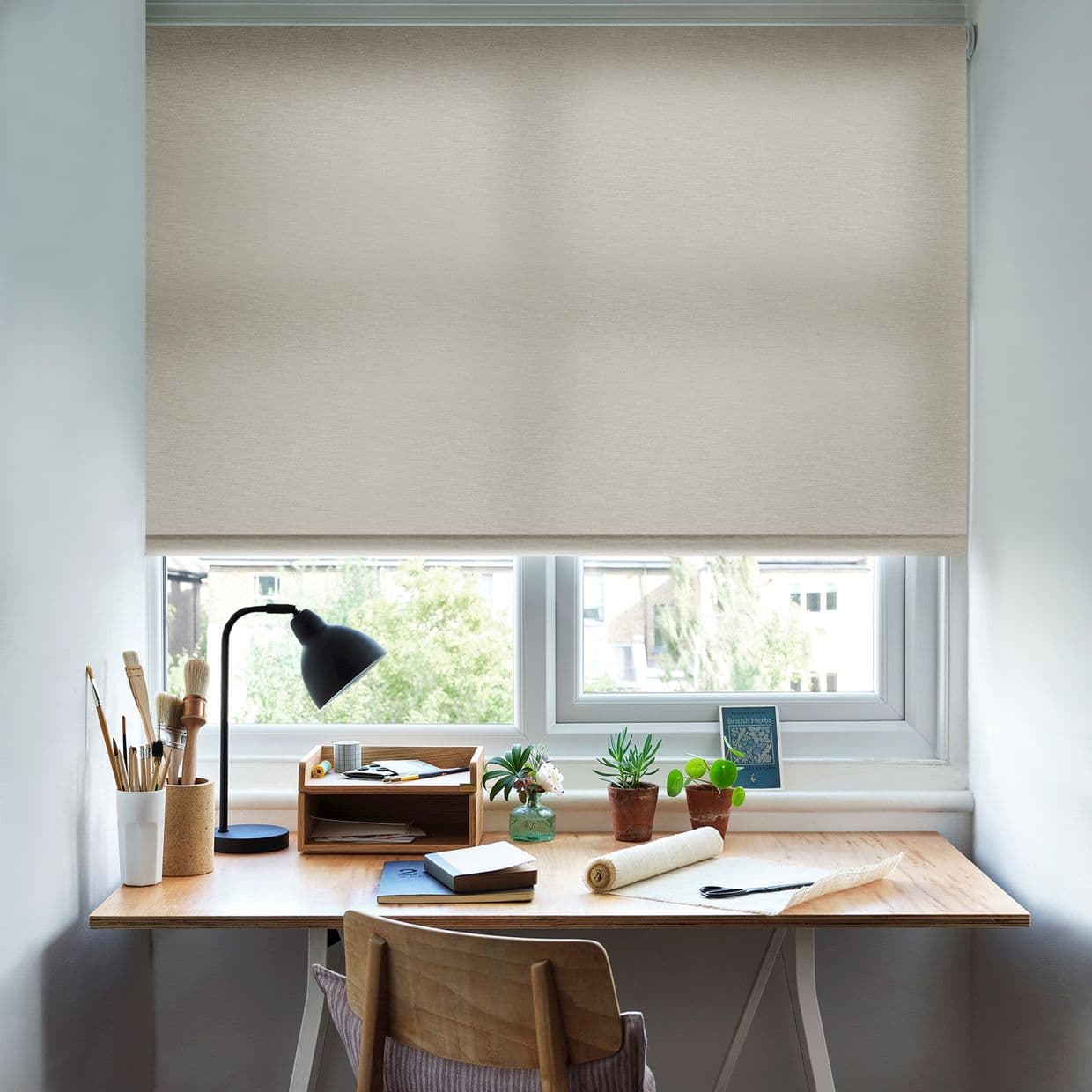 Beige fabric roller blind, lowered nearly fully and softly filtering daylight; overlooking a small home-office window. Wooden desk beneath holds a black desk lamp, brushes, notebooks and potted succulents.
