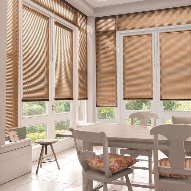 Beige horizontal slatted blinds on tall French doors and windows, lowered with slats mostly closed, filtering warm daylight into a bright dining area with white table, chairs and garden view.