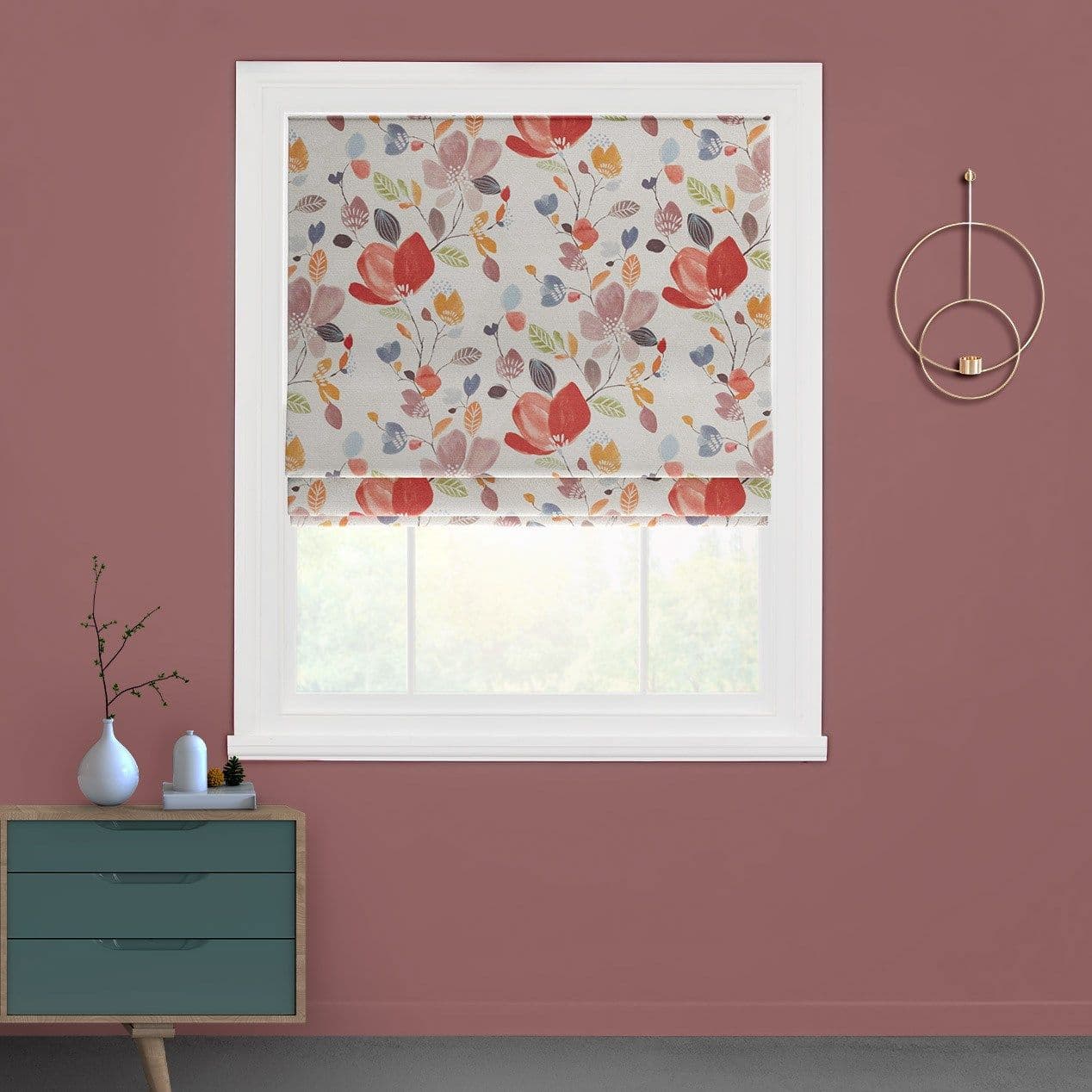 Floral Roman blind, partially lowered in soft folds, filtering daylight through a white-framed window in a mauve-walled room with a teal chest and circular wall sconce; blurred greenery outside.