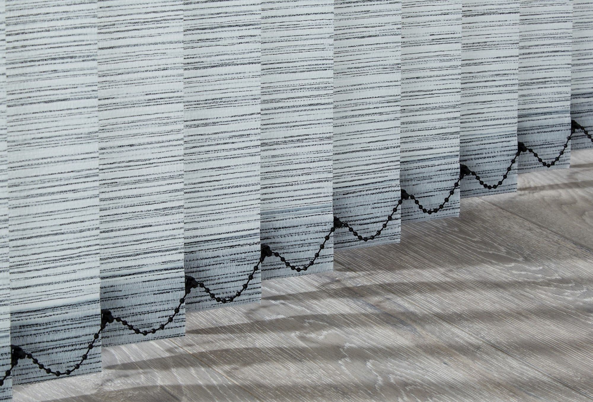Vertical fabric blinds: light-gray slats with horizontal black stripes and a scalloped black bead motif along lower edges â€” hanging closed â€” filtering daylight, casting shadows on a light wood floor.