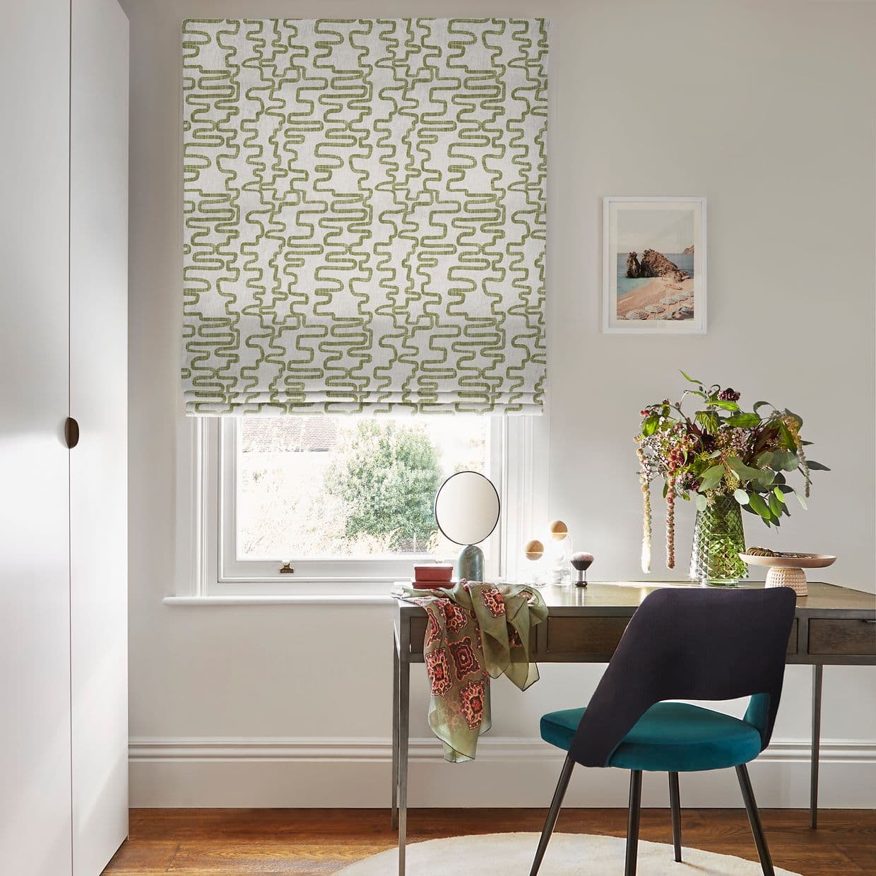 A green-squiggle patterned Roman blind covers the window, lowered and softly filtering daylight; a home study desk beneath holds a round mirror, vase of foliage, teal chair, and framed coastal print.