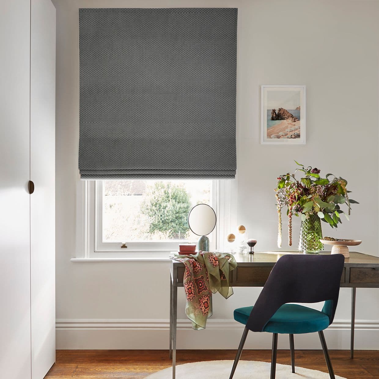 Gray patterned Roman blind lowered over a sash window, leaving a small gap at the bottom. Bright home study with a desk, teal chair and a vase of flowers.