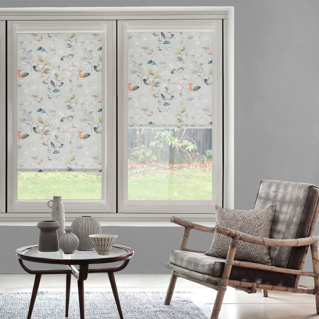 Patterned roller blinds with pastel watercolor leaf motif covering twin windowsâ€”left fully lowered, right partially raisedâ€”diffusing daylight into a grey living room with rattan leather armchair, table and backyard view.