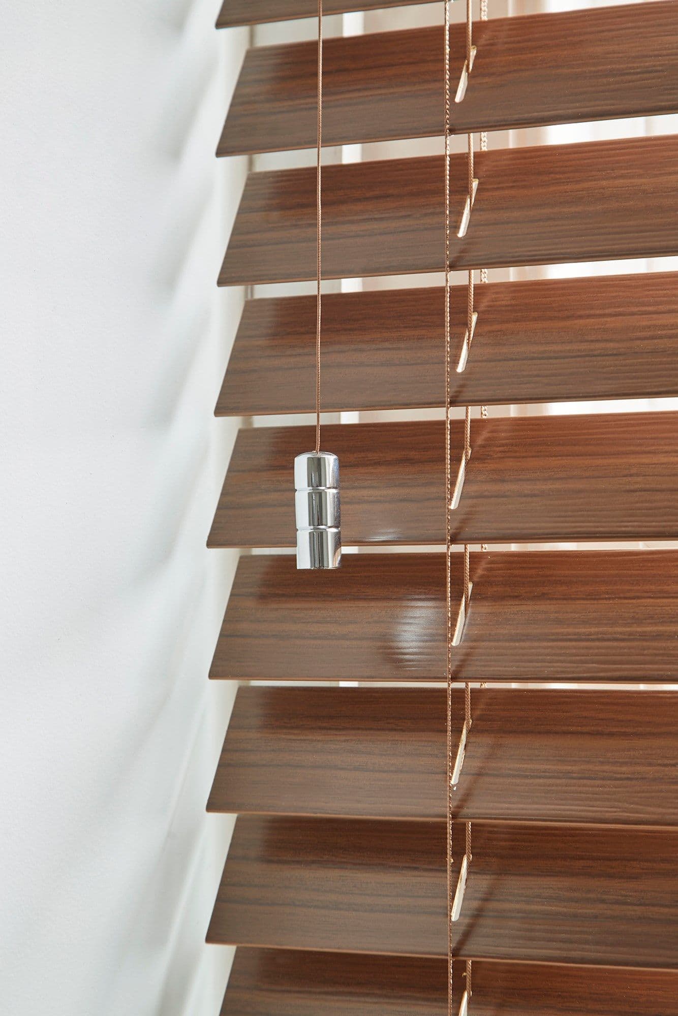 Wooden horizontal venetian blinds with brown wood-grain slats and pull cords bearing a chrome weight; partially closed, slightly angled to filter light in a close-up against a white wall.