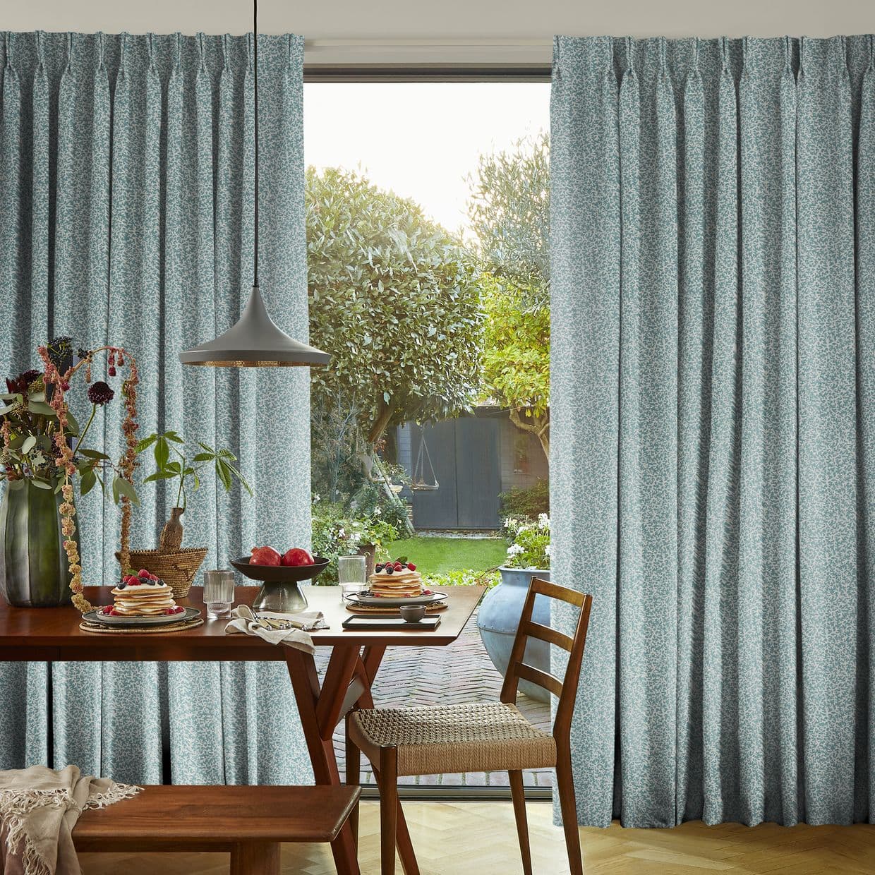 Light-blue, pleated patterned curtains hang at a sliding glass door, drawn slightly open to let daylight in, framing a dining table set and a lush garden view.