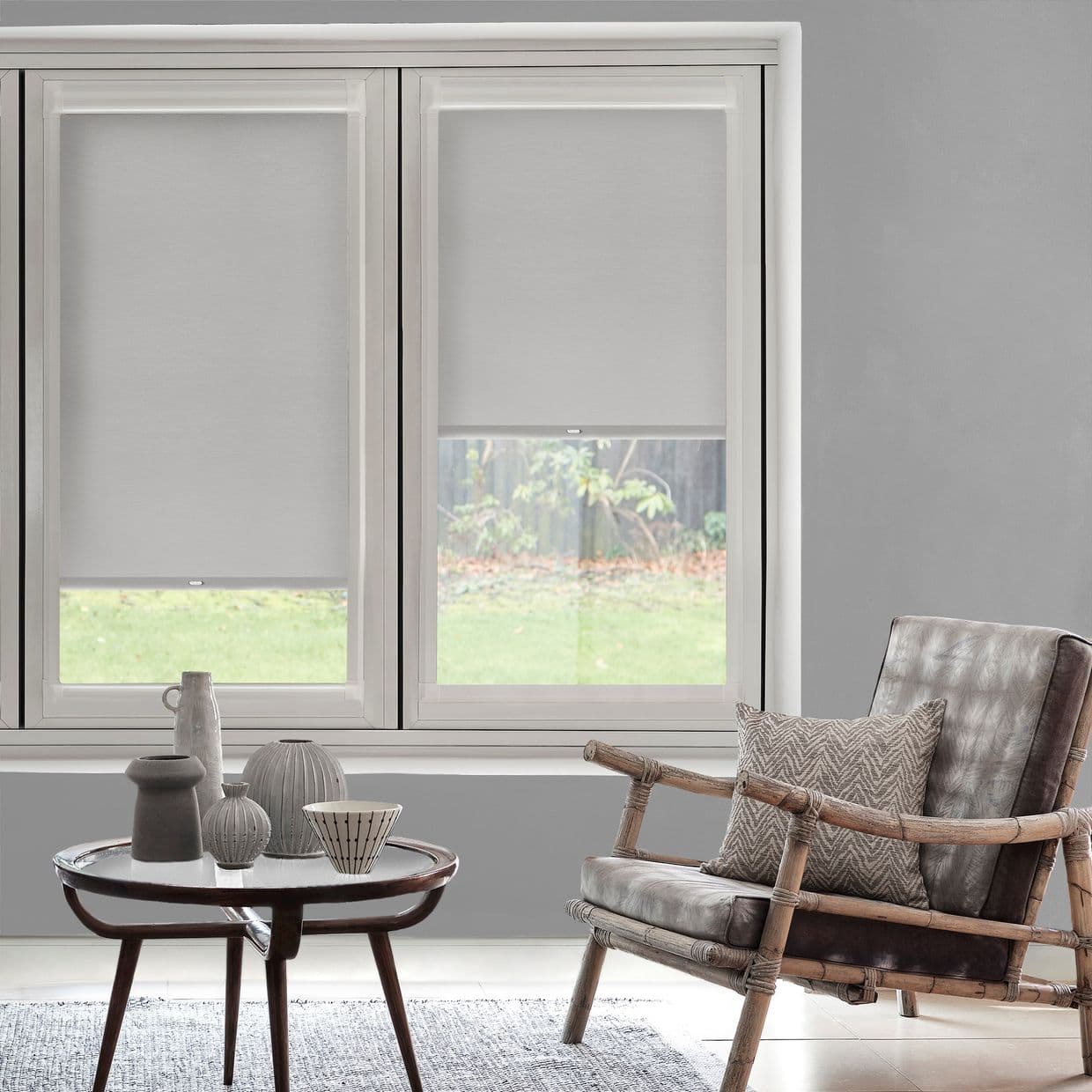 Gray roller blinds, fitted over two adjacent windows and partially lowered to reveal narrow garden views, filtering daylight; above a living room with rattan armchair, cushion and small table.