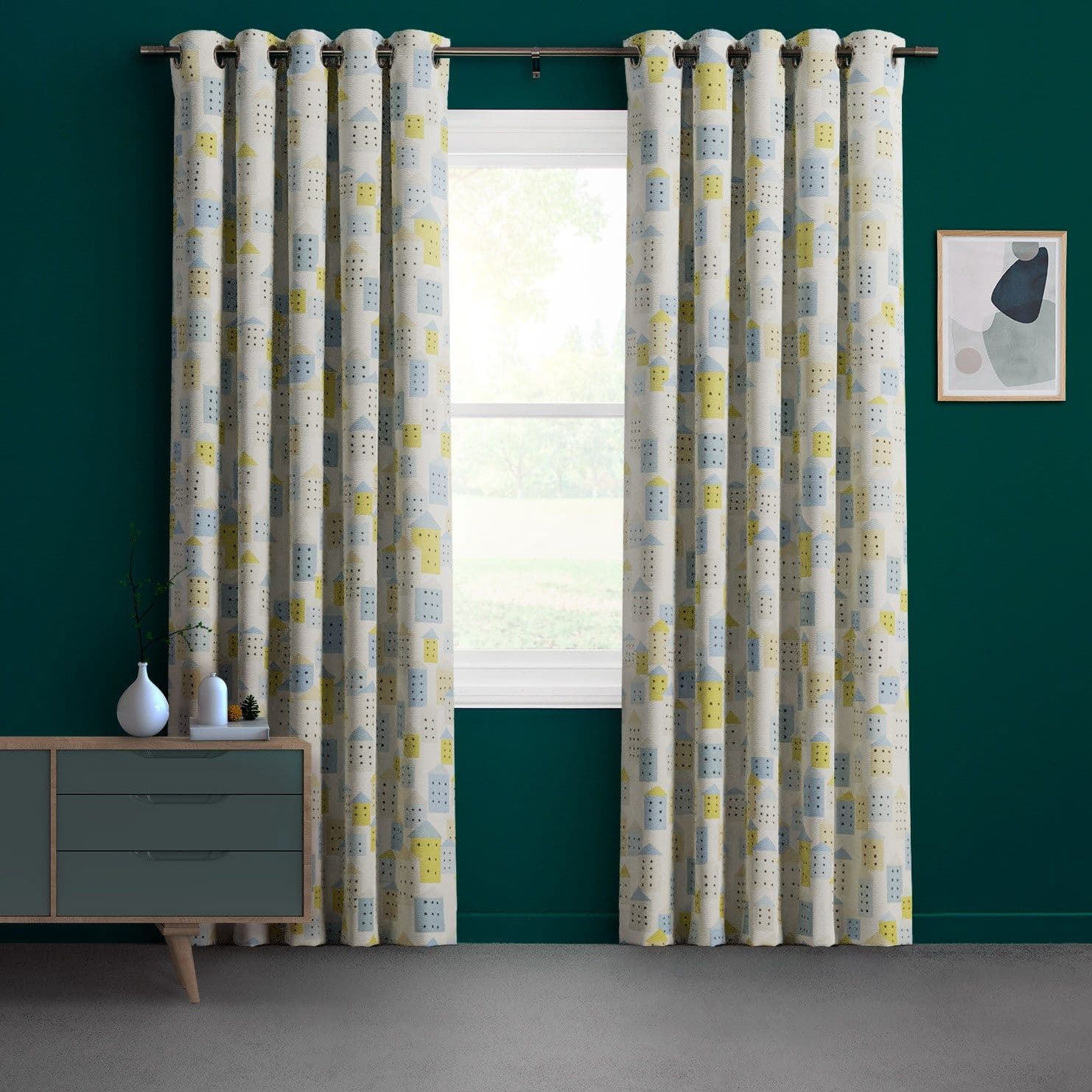 Grommet-top patterned curtains, cream with blue-yellow geometric house motifs, hung in two panels on a metal rod, drawn aside to reveal a sunlit window â€” teal-walled living room.
