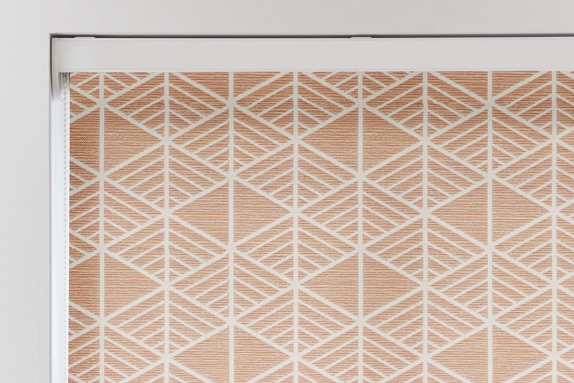 A fabric roller blind with white geometric diamond lattice over peach woven texture, mounted in a white cassette and lowered (closed), filtering light in a bright, neutral interior window recess.