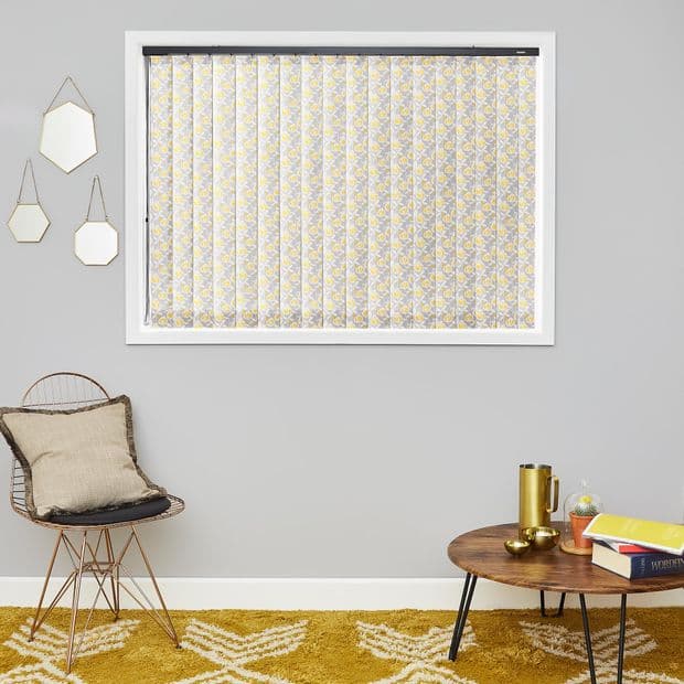 Patterned vertical blinds (yellow and gray floral) cover the window, closed and filtering light on a gray wall in a living room with chair, wooden coffee table and three mirrors.