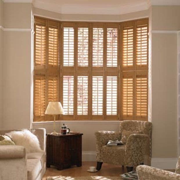 Wooden plantation shutters span a multi-panel bay window, slats partially angled to filter warm daylight; living room setting with upholstered chairs, side table, lamp and soft neutral decor.