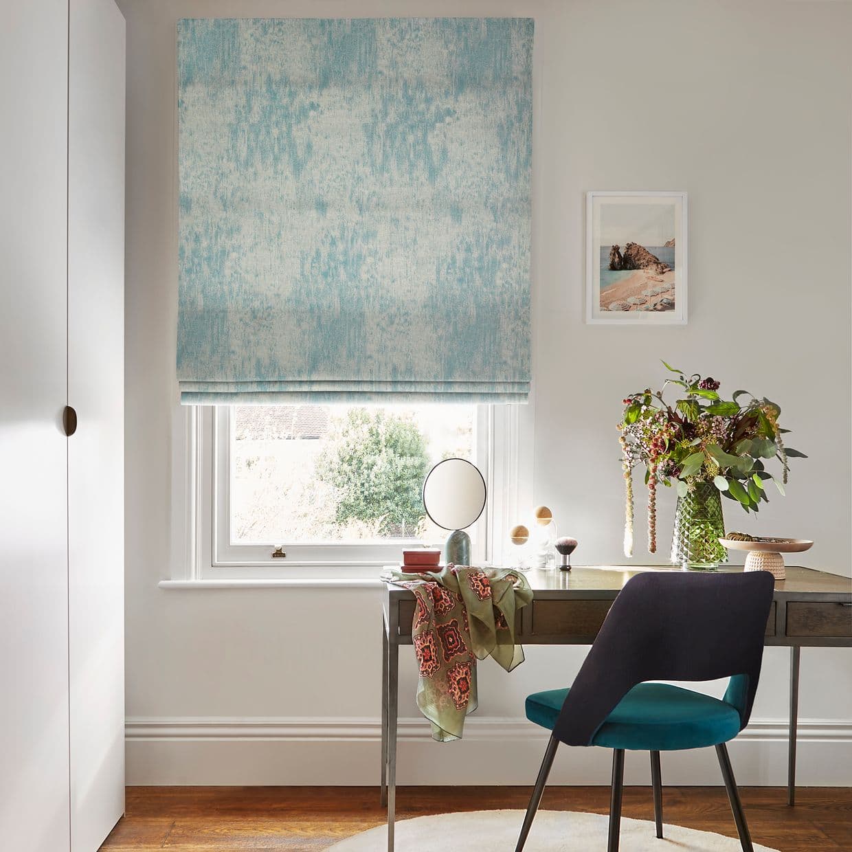 Turquoise textured roman blind lowered over a window, softly filtering daylight. A desk with round mirror, vase of foliage and teal chair sits in a bright home study.