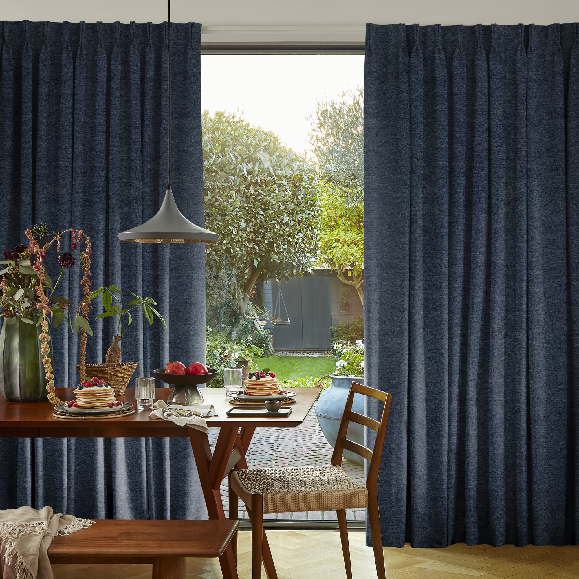 Navy pleated floor-length curtains, parted at center; curtains framing and filtering soft daylight onto a wooden table set for breakfast in a cozy nook opening to a sunlit garden.