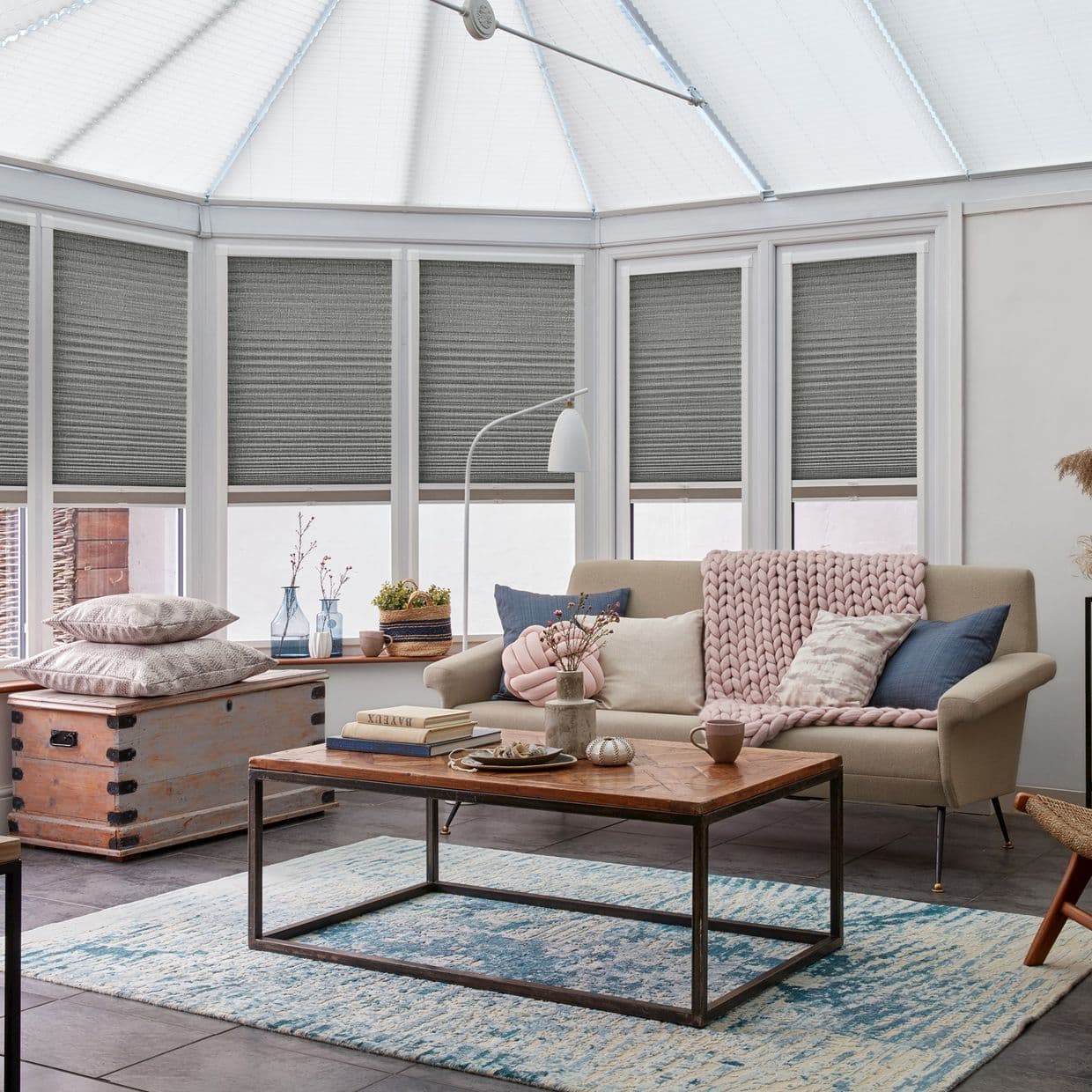 Gray textured cellular blinds, lowered to mid-window across a conservatory bay, partially filtering daylight; in a sitting room with beige sofa, pink chunky throw, wooden coffee table and blue rug.