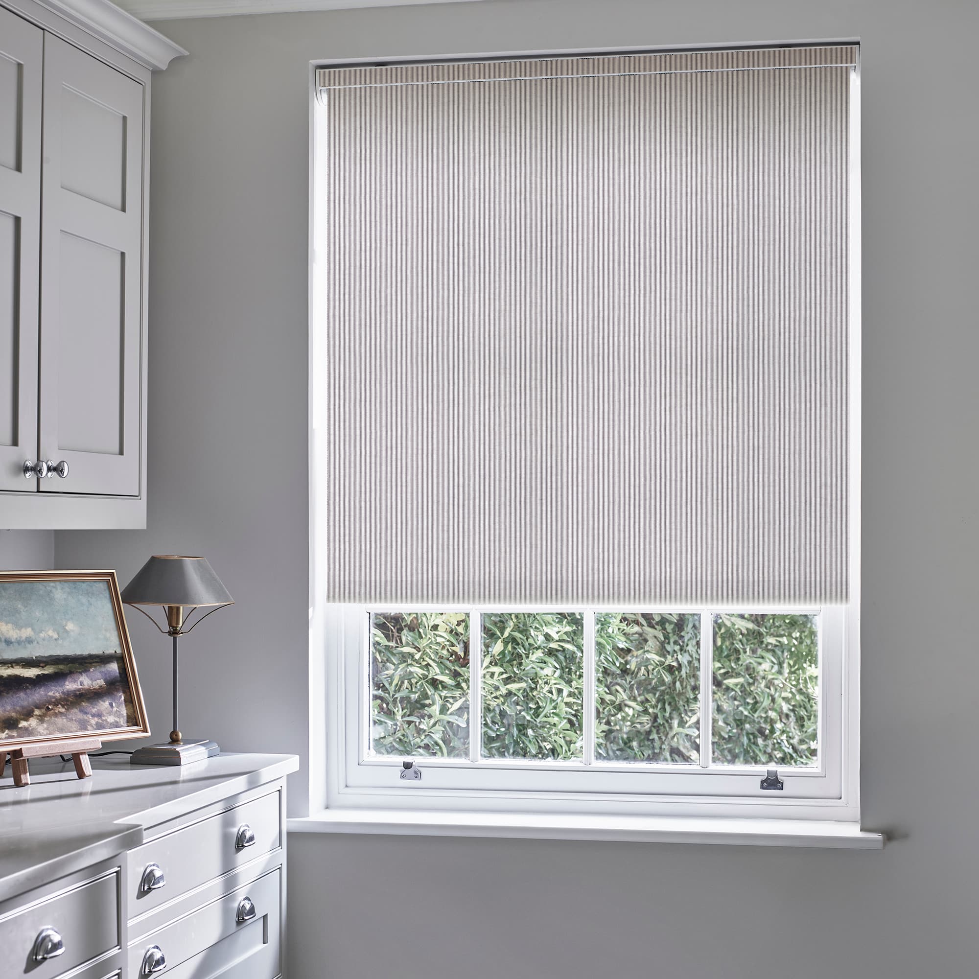 Striped pleated roller blind, lowered nearly to the sill with thin vertical pinstripes; blocking and softly diffusing daylight over a pale-grey room with white cabinetry, lamp, and leafy outside view.