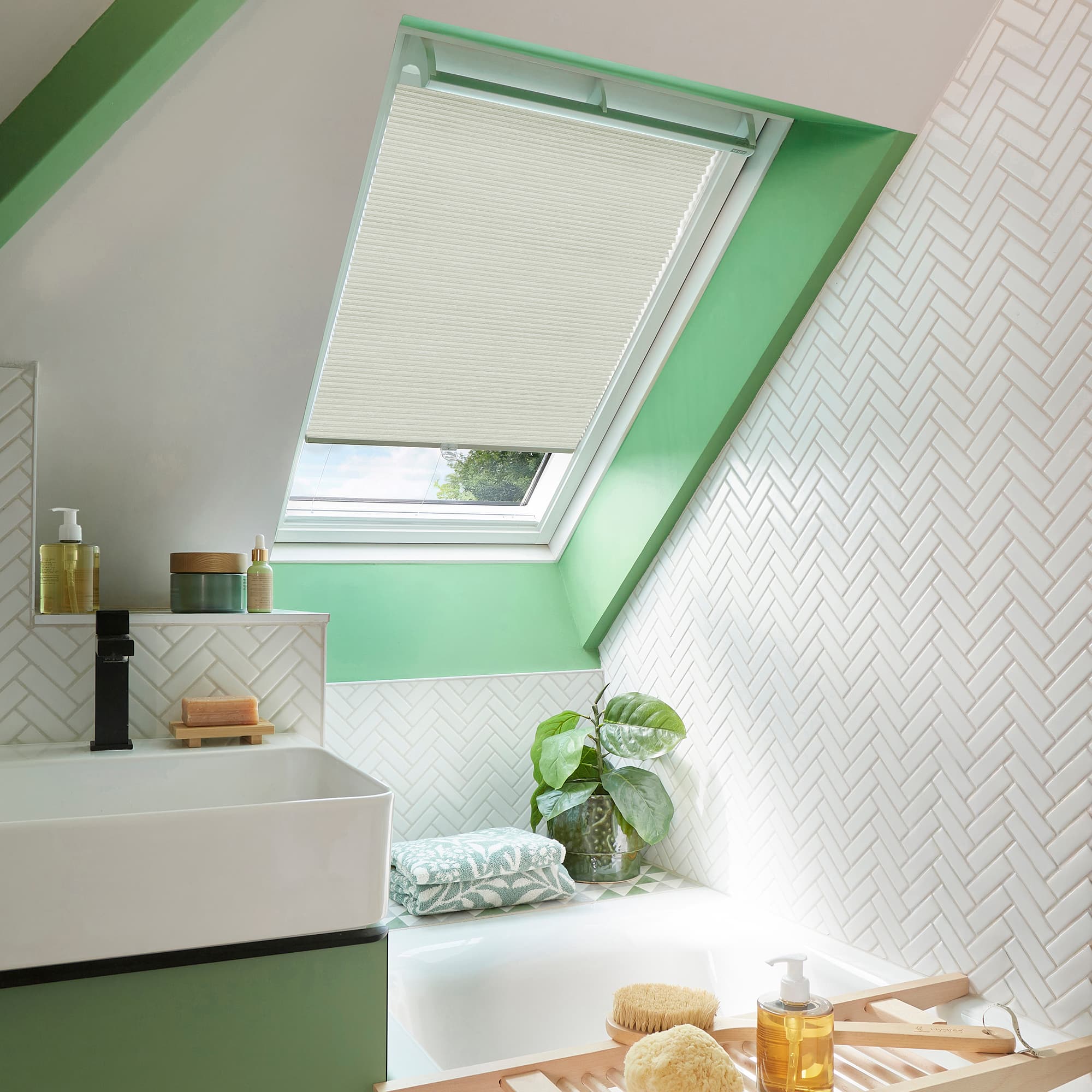 Cream pleated skylight blind, lowered over a sloped roof window, filtering soft daylight into a bright attic bathroom with green trim, white herringbone tiles, sink, tub and plant.