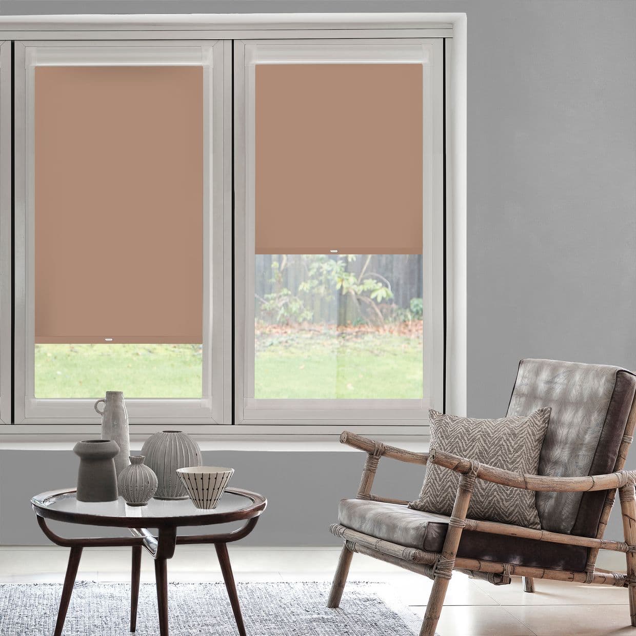 Two terracotta roller blinds cover twin windowsâ€”left fully lowered, right halfwayâ€”partially blocking and diffusing light in a neutral living room with a rattan armchair, coffee table, grassy backyard view.