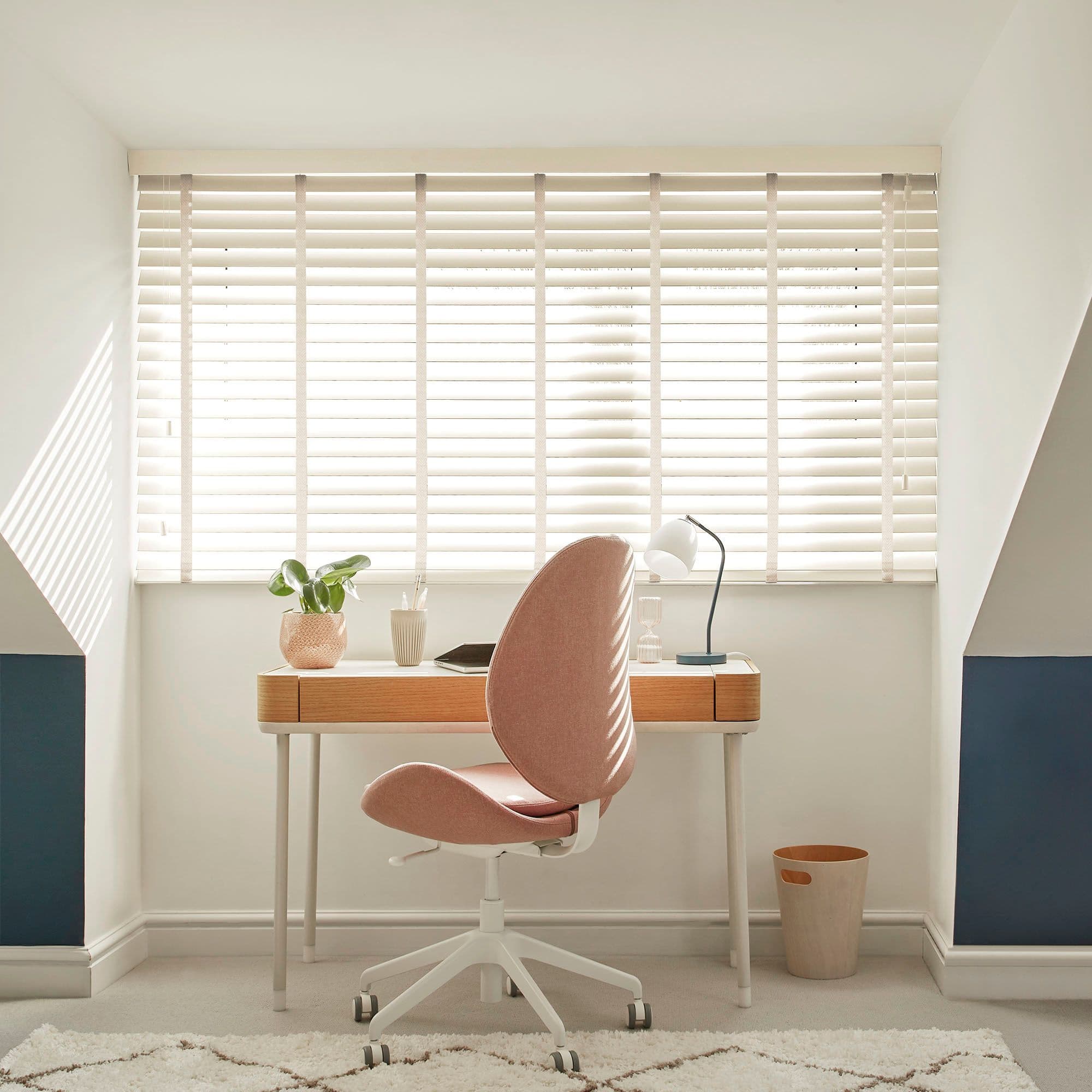 White horizontal Venetian blinds across a wide window, slats tilted with fabric tapes; filtering bright daylight and casting striped shadows; attic home-office niche with wooden desk, pink swivel chair, lamp.