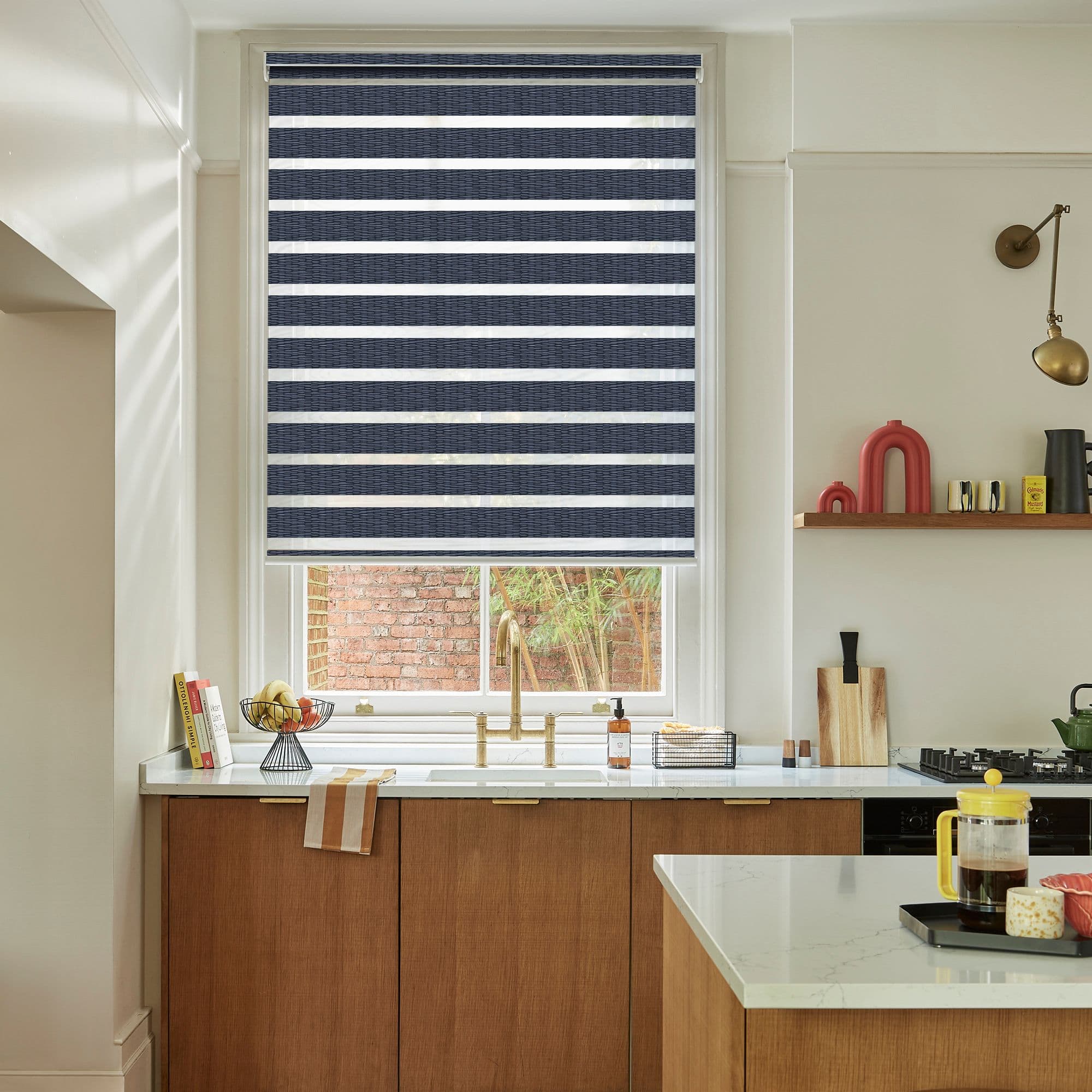 Dual-layer zebra roller blind with alternating navy textured and sheer white horizontal stripes, lowered and filtering light, above a kitchen sink with brass faucet, wooden cabinets, and marble countertops.
