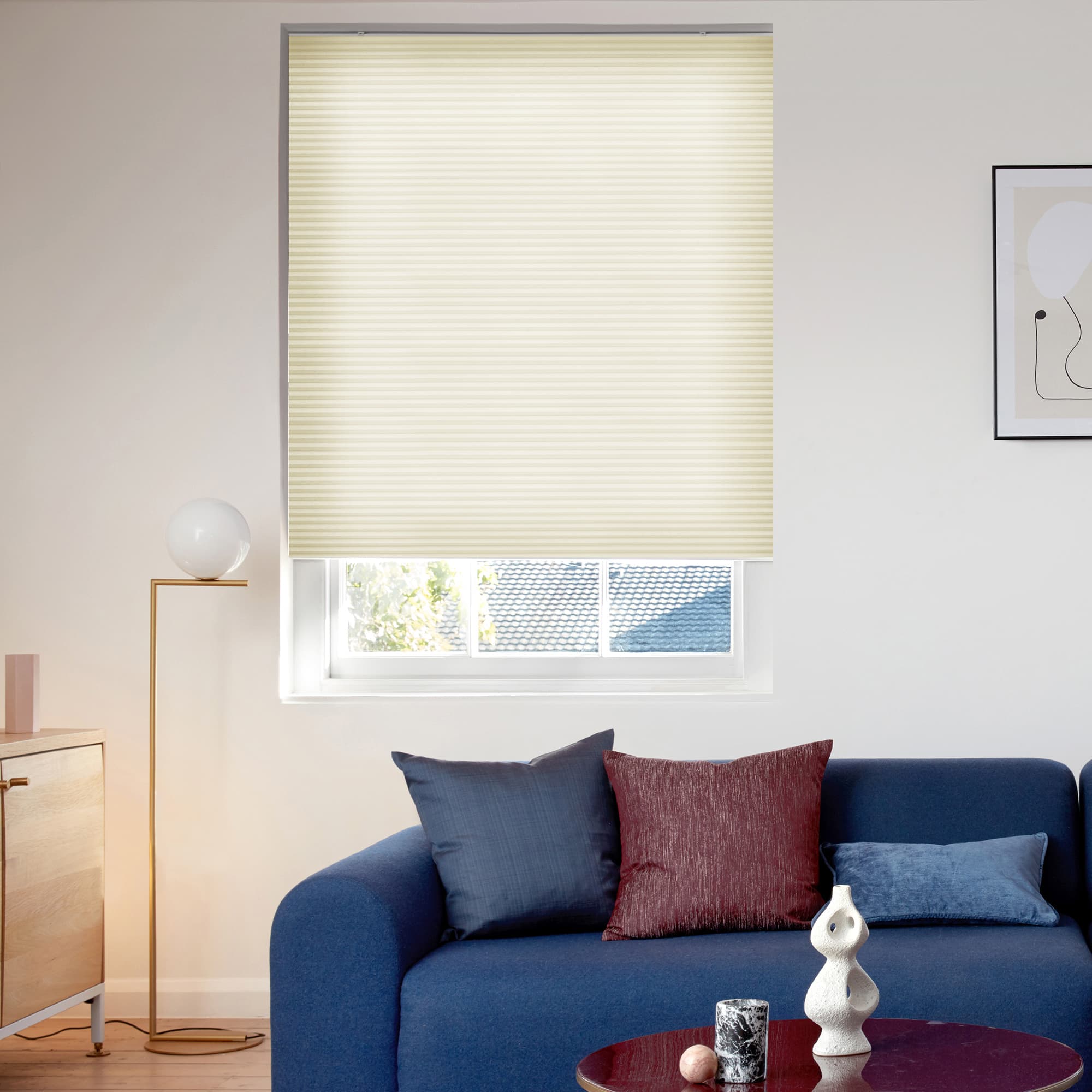 Cream pleated blind, lowered to cover most of the window with horizontal ribs, filtering soft daylight; above window in a bright living room with a blue sofa and a lamp.