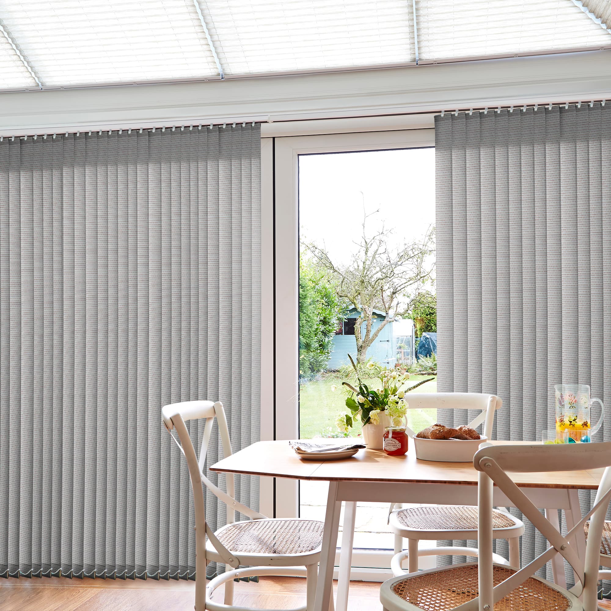 Vertical fabric blinds, light gray, partly parted to reveal a central glass patio door, slats hanging straight; filtering soft daylight into a dining nook with wooden table, white chairs, garden view.