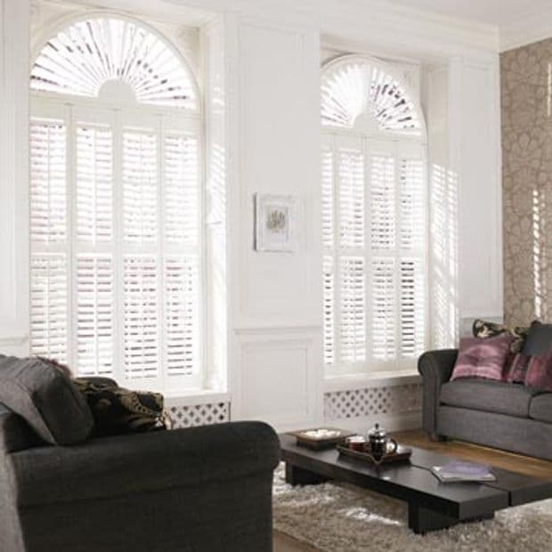 White plantation shutters cover tall arched windowsâ€”rectangular louvered panels below and fan-shaped radial topsâ€”closed, filtering sunlight and casting patterned shadows in a living room with sofas, coffee table, and rug.