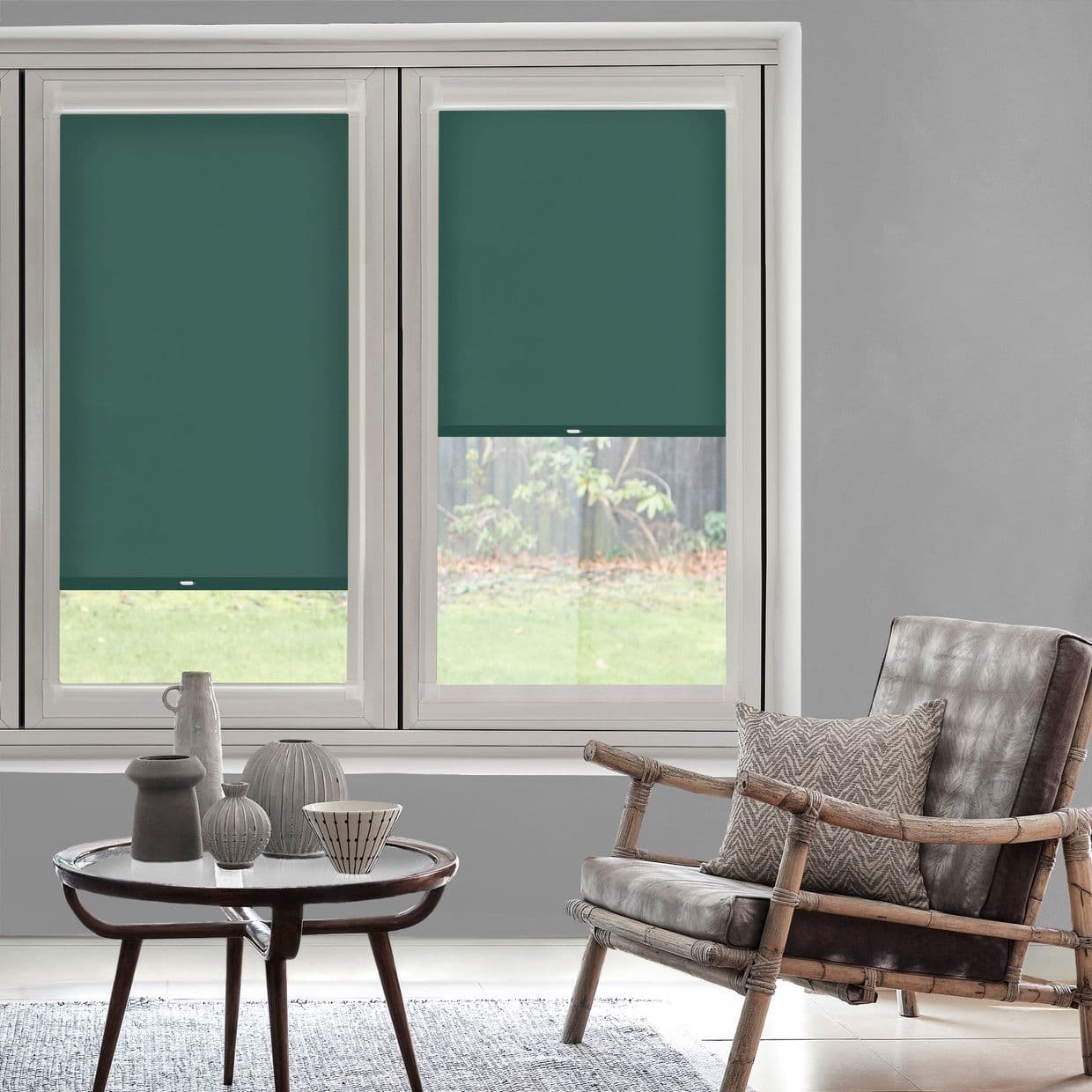 Teal roller blinds, two panels â€” left fully lowered, right partially lowered with pull tab â€” blocking light over window in a softly lit living room with a rattan armchair and table.