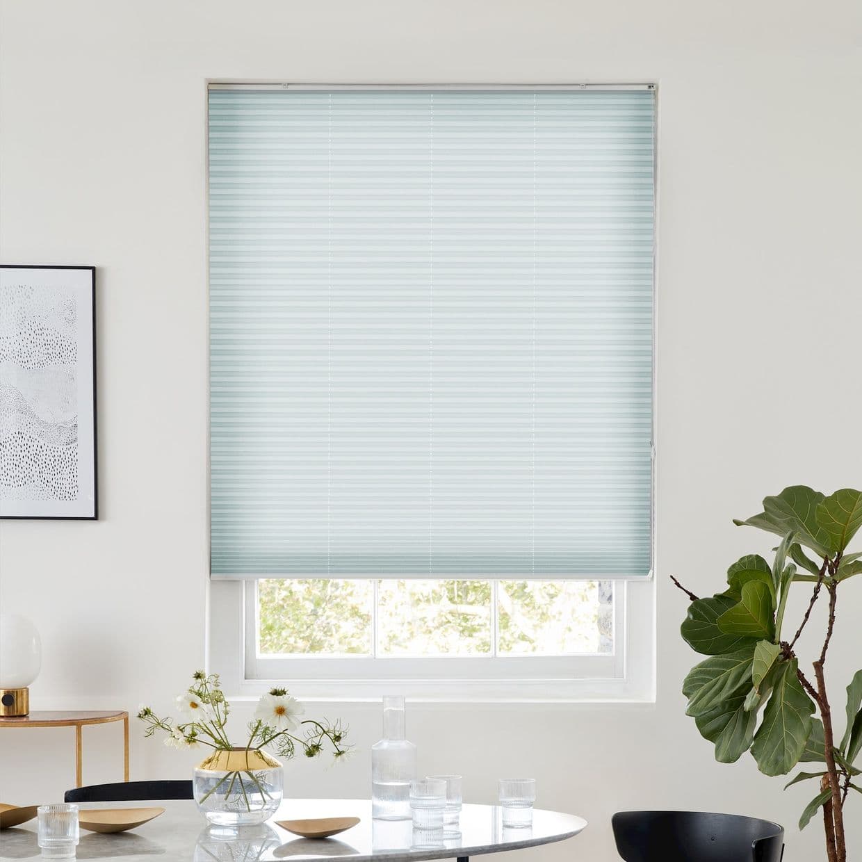 Pale blue pleated blind lowered, filtering soft daylight; white-framed window with lower sash visible. Dining area: round table with vase and glassware, framed art left, potted plant right.